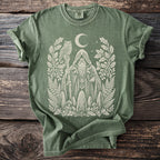 Green t-shirt with cream print of a hooded figure, owl, moon, and forest elements.