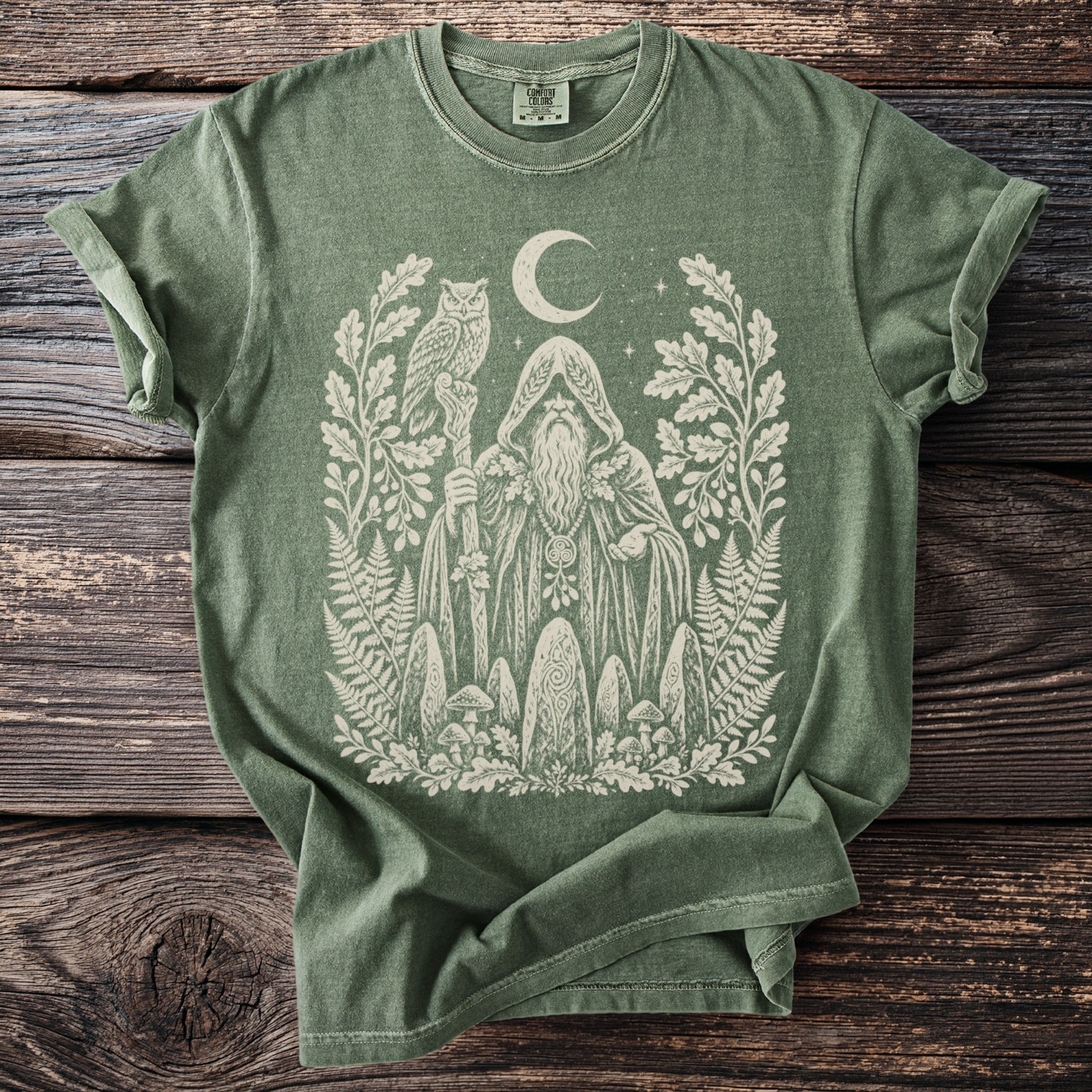 Green t-shirt with cream print of a hooded figure, owl, moon, and forest elements.