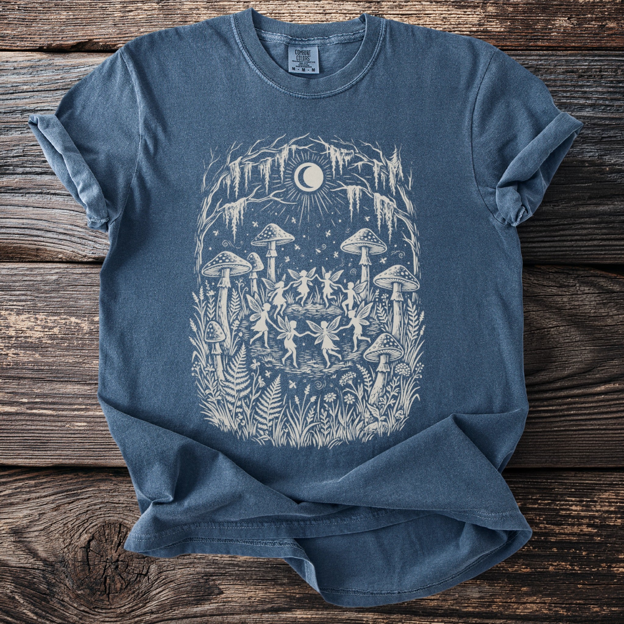 Blue t-shirt with a graphic of fairies dancing around mushrooms under a crescent moon.