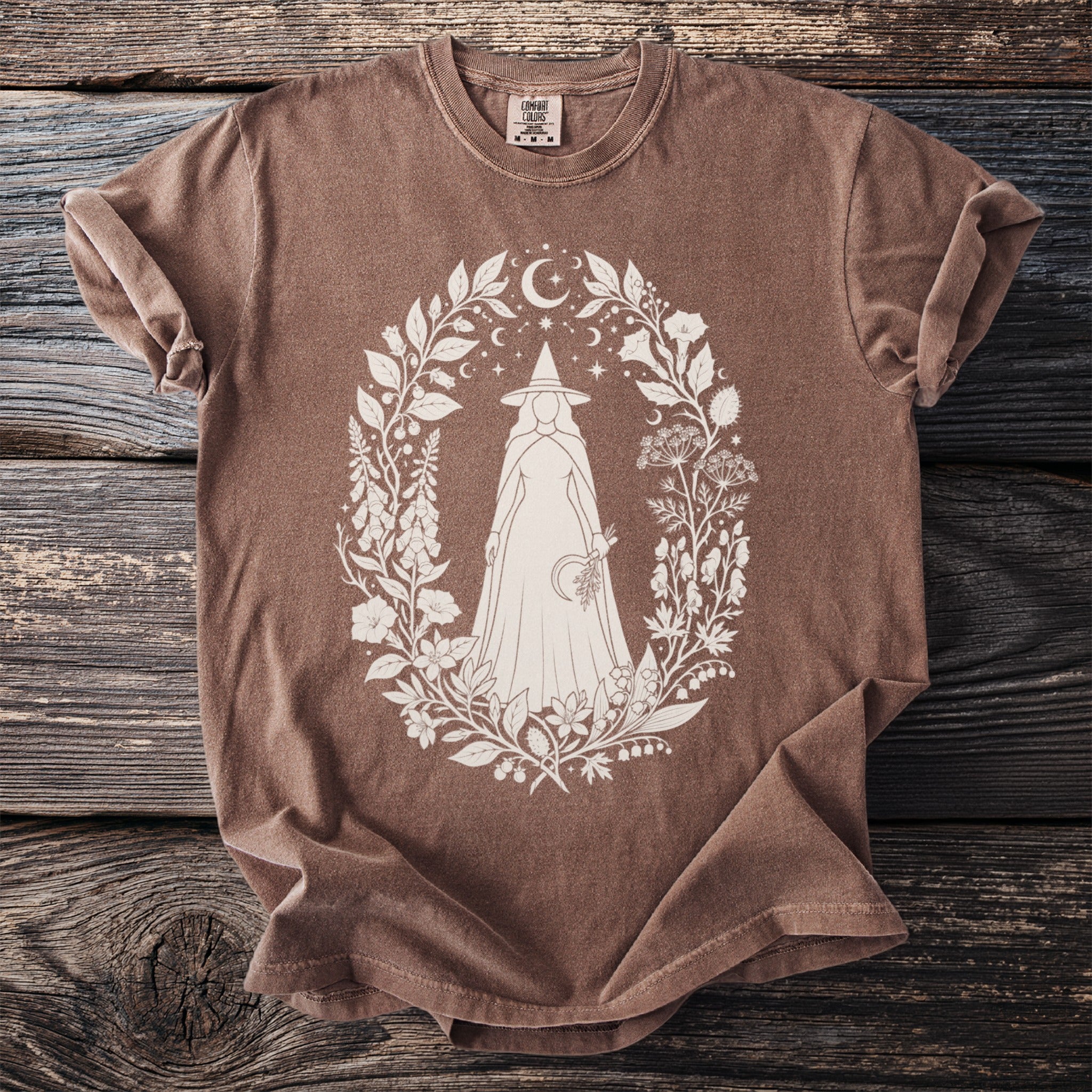 Brown t-shirt with white graphic of a cloaked figure within a floral and celestial wreath.