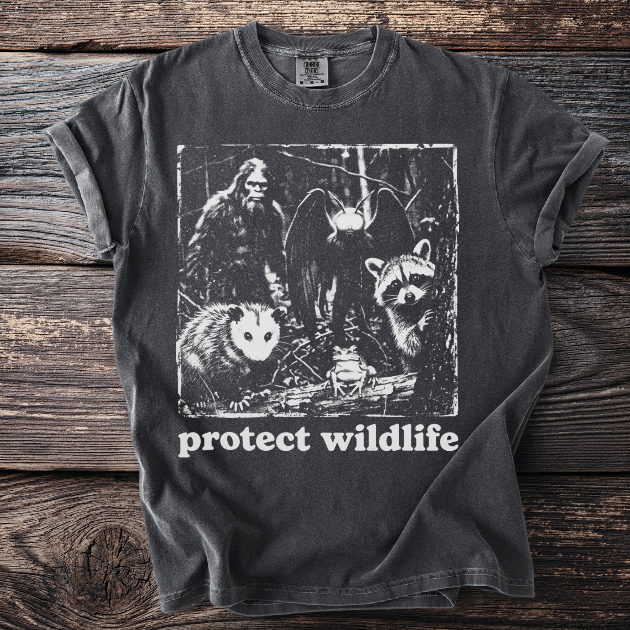 Dark t-shirt with Bigfoot, moth, opossum, frog, and raccoon graphic and "protect wildlife" text.