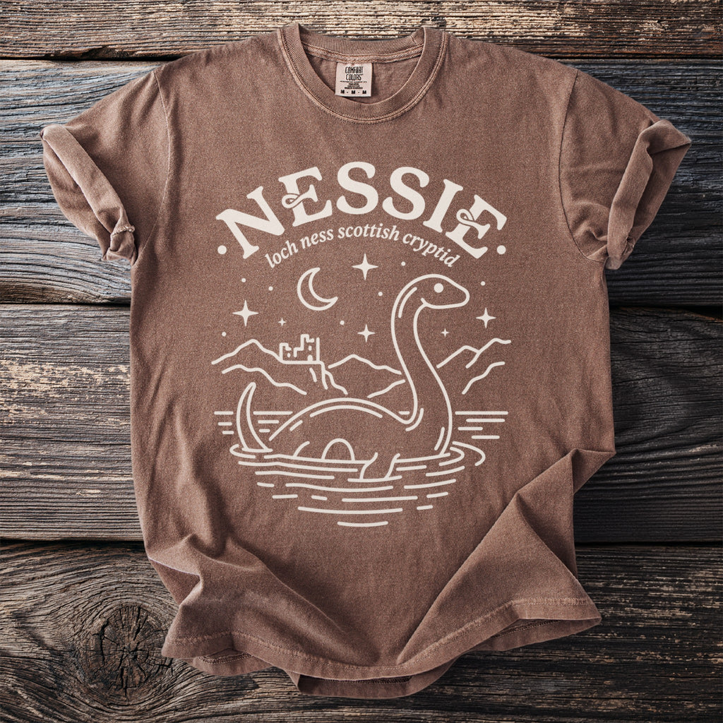 Brown t-shirt with white graphic of Nessie, castle, moon, and stars