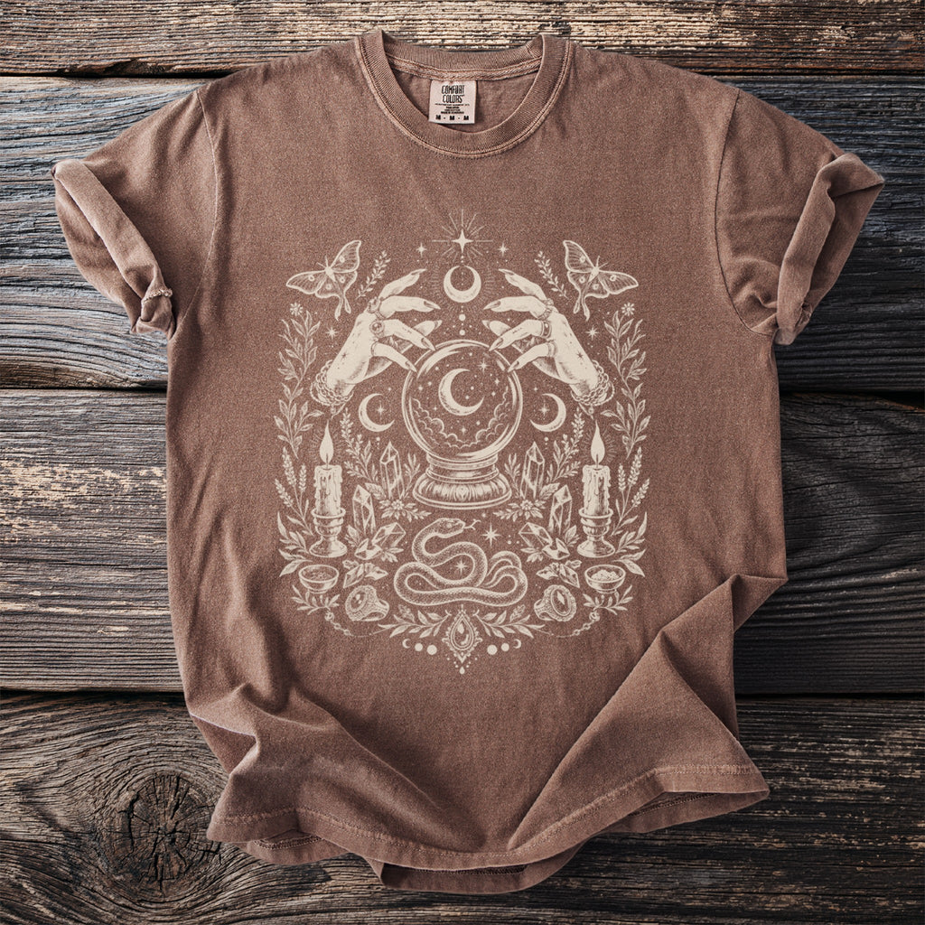 Brown t-shirt with cream graphic featuring hands, crystal ball, moth, candles, crystals, and snake.