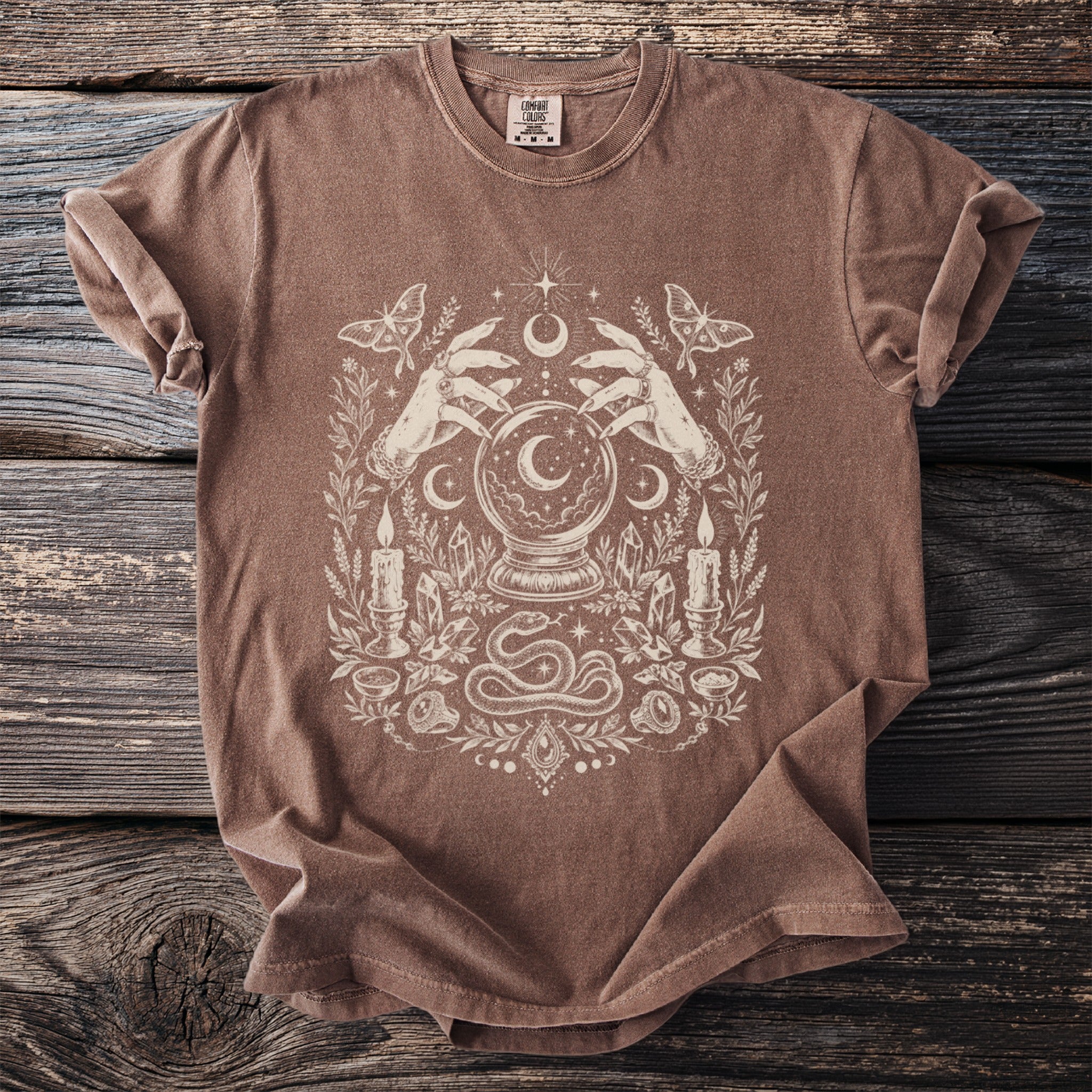 Brown t-shirt with cream graphic featuring hands, crystal ball, moth, candles, crystals, and snake.