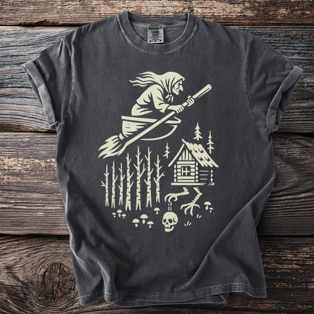 Dark t-shirt with a creamy white graphic of a witch flying a broom over a forest cabin.