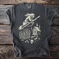 Dark t-shirt with a creamy white graphic of a witch flying a broom over a forest cabin.