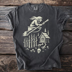 Dark t-shirt with a creamy white graphic of a witch flying a broom over a forest cabin.