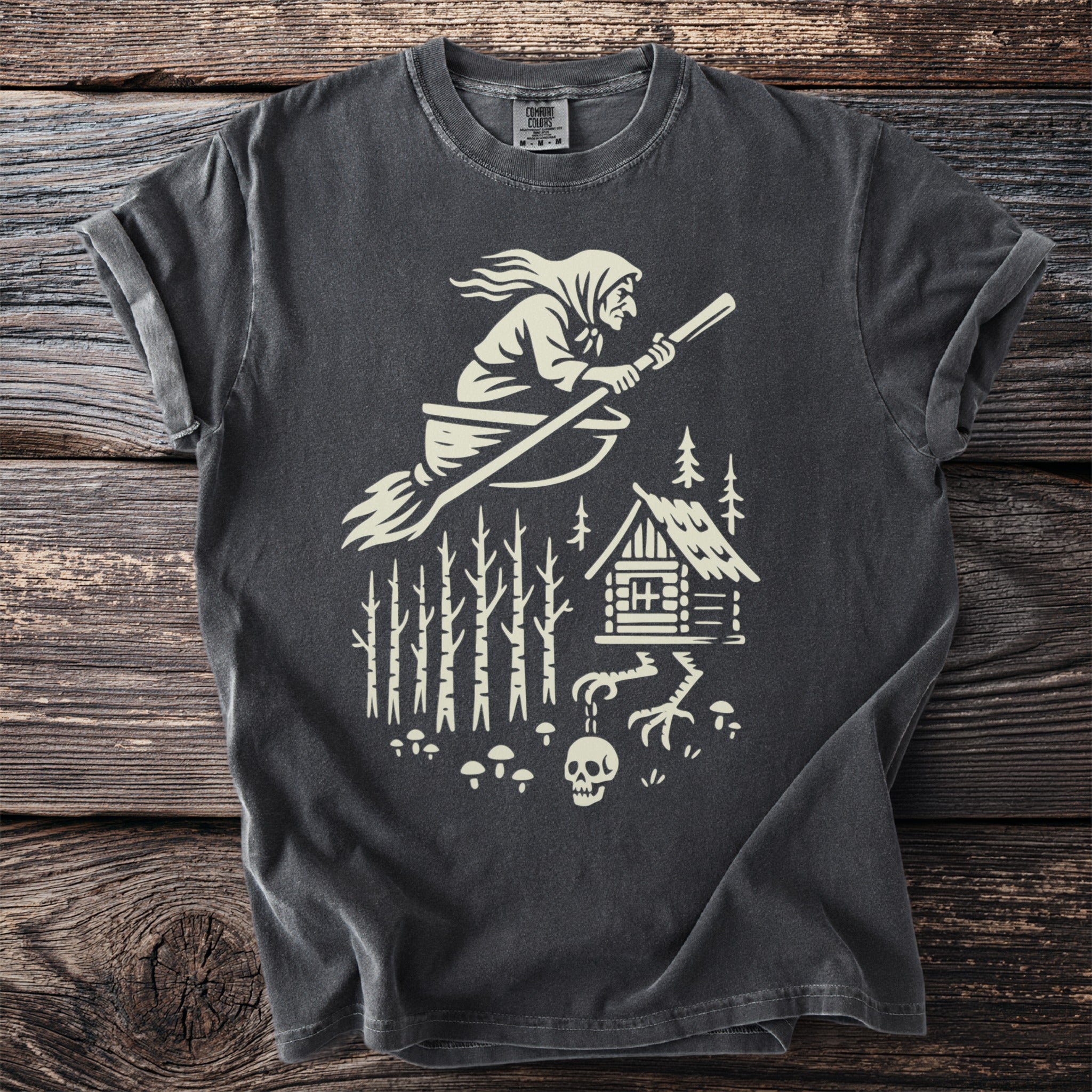 Dark t-shirt with a creamy white graphic of a witch flying a broom over a forest cabin.