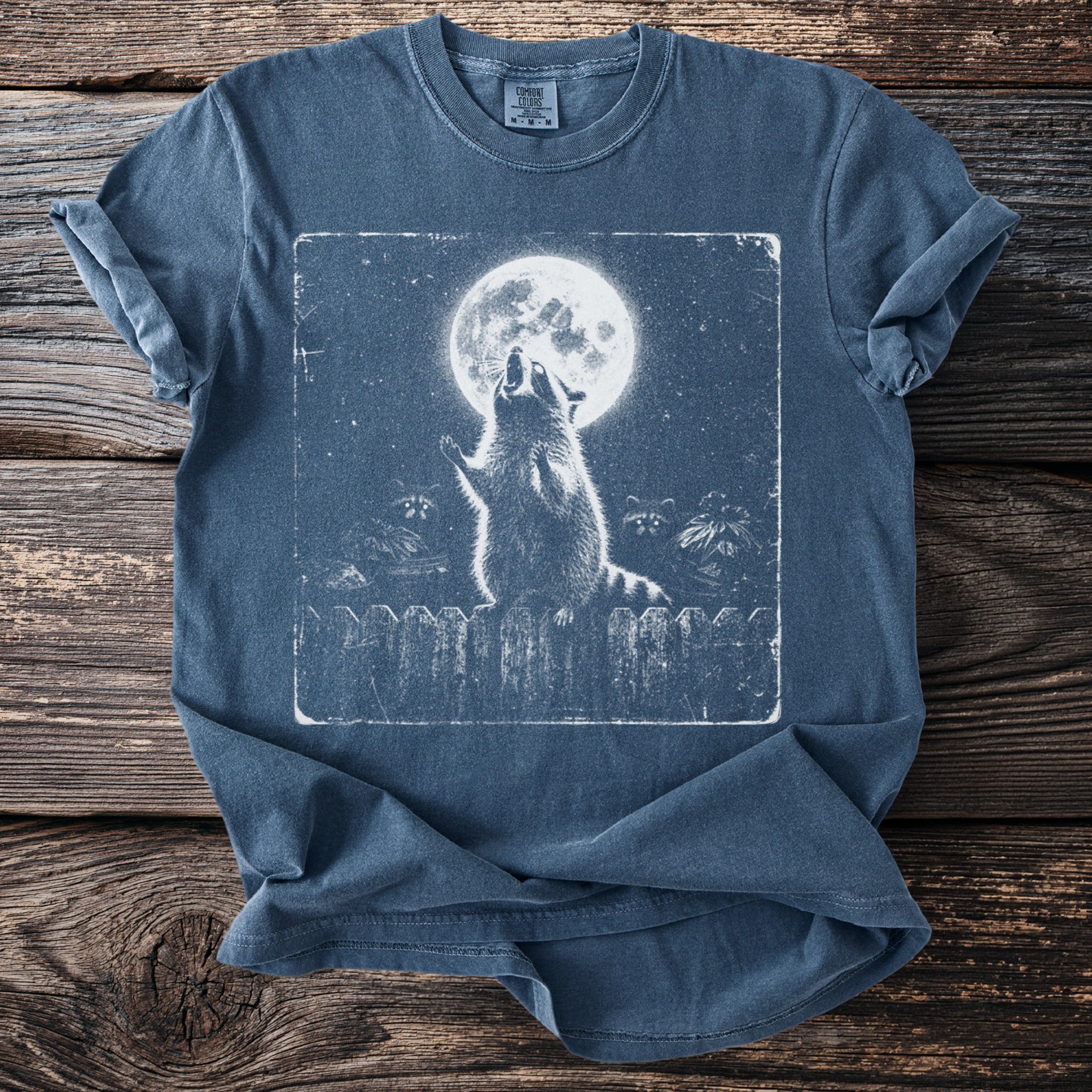 Blue t-shirt with a graphic of raccoons howling at the moon on a fence.