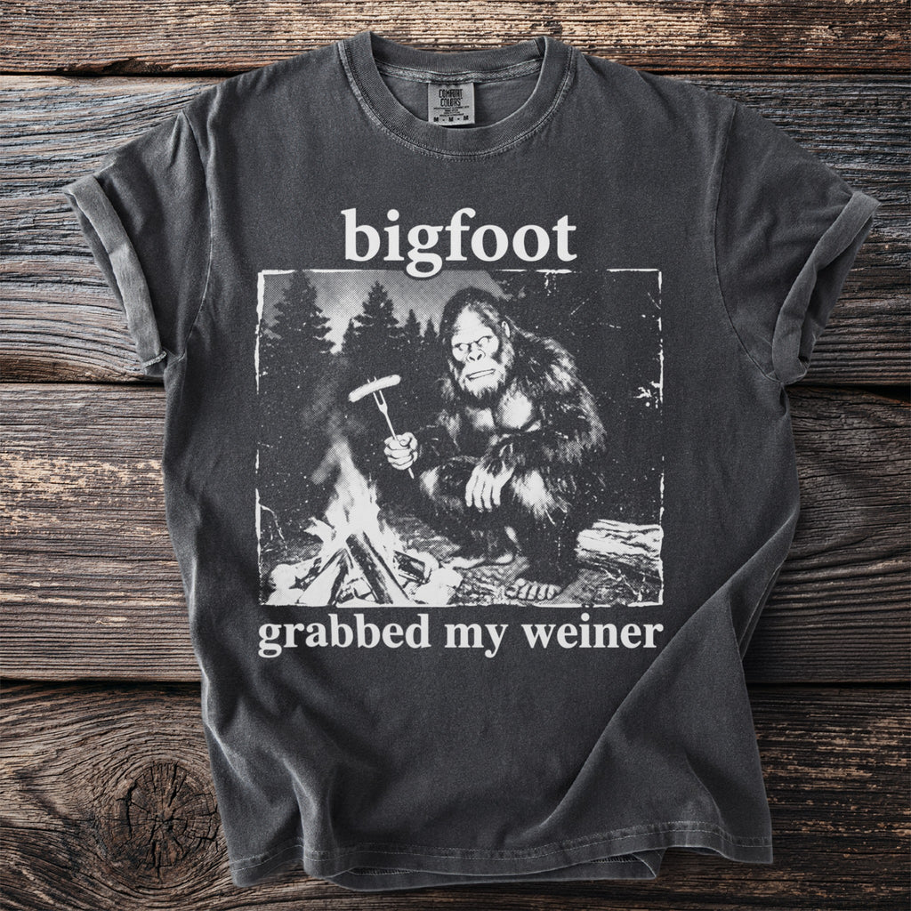 Bigfoot sitting by a campfire holding a sausage on a fork, t-shirt graphic.
