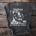 Bigfoot sitting by a campfire holding a sausage on a fork, t-shirt graphic.