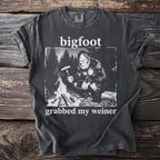 Bigfoot sitting by a campfire holding a sausage on a fork, t-shirt graphic.