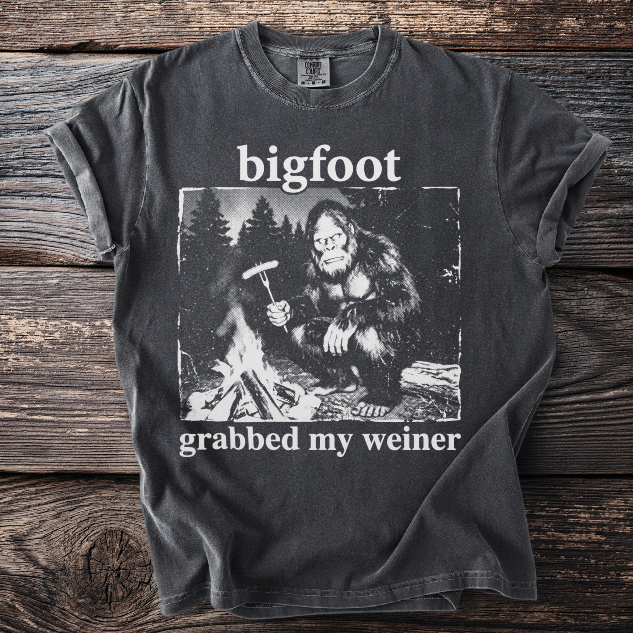 Bigfoot sitting by a campfire holding a sausage on a fork, t-shirt graphic.