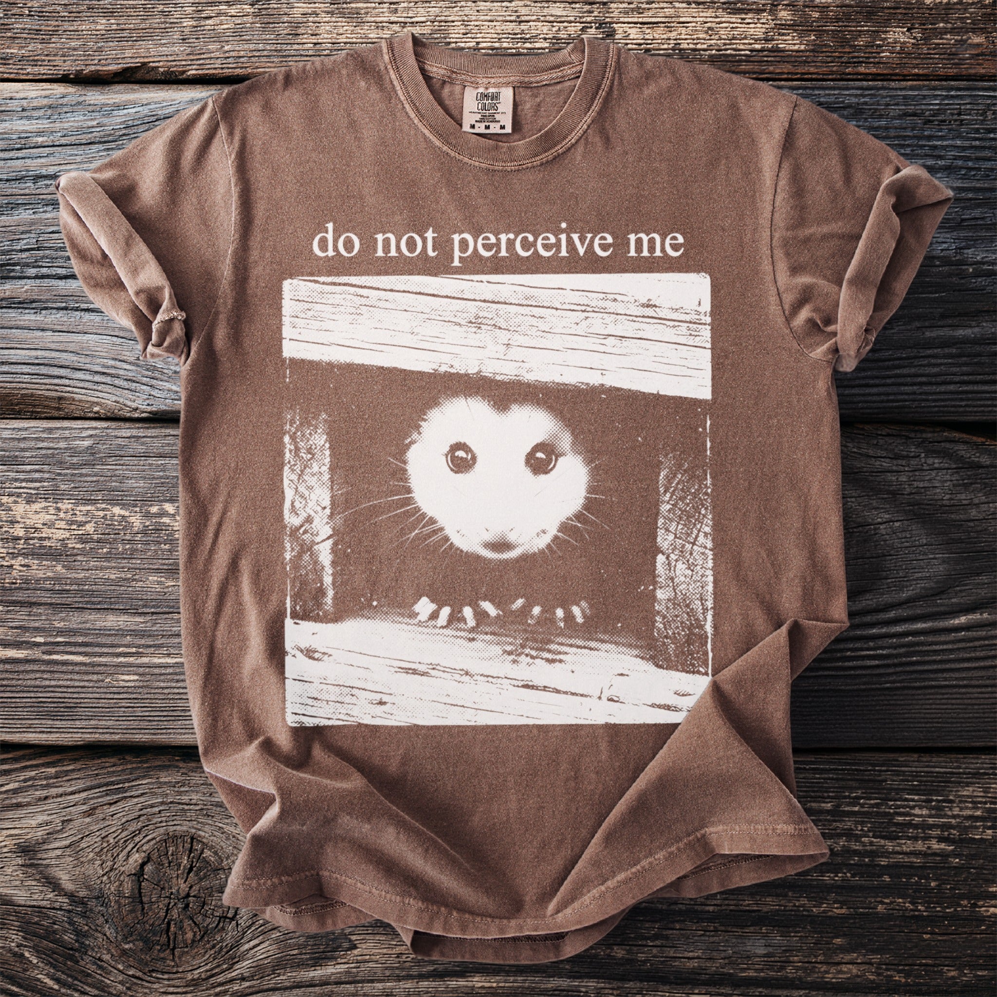 Brown t-shirt graphic of a possum peeking out of a box, with text "do not perceive me".