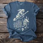 Blue t-shirt with a cream graphic of a witch flying on a broom over a cabin and forest.