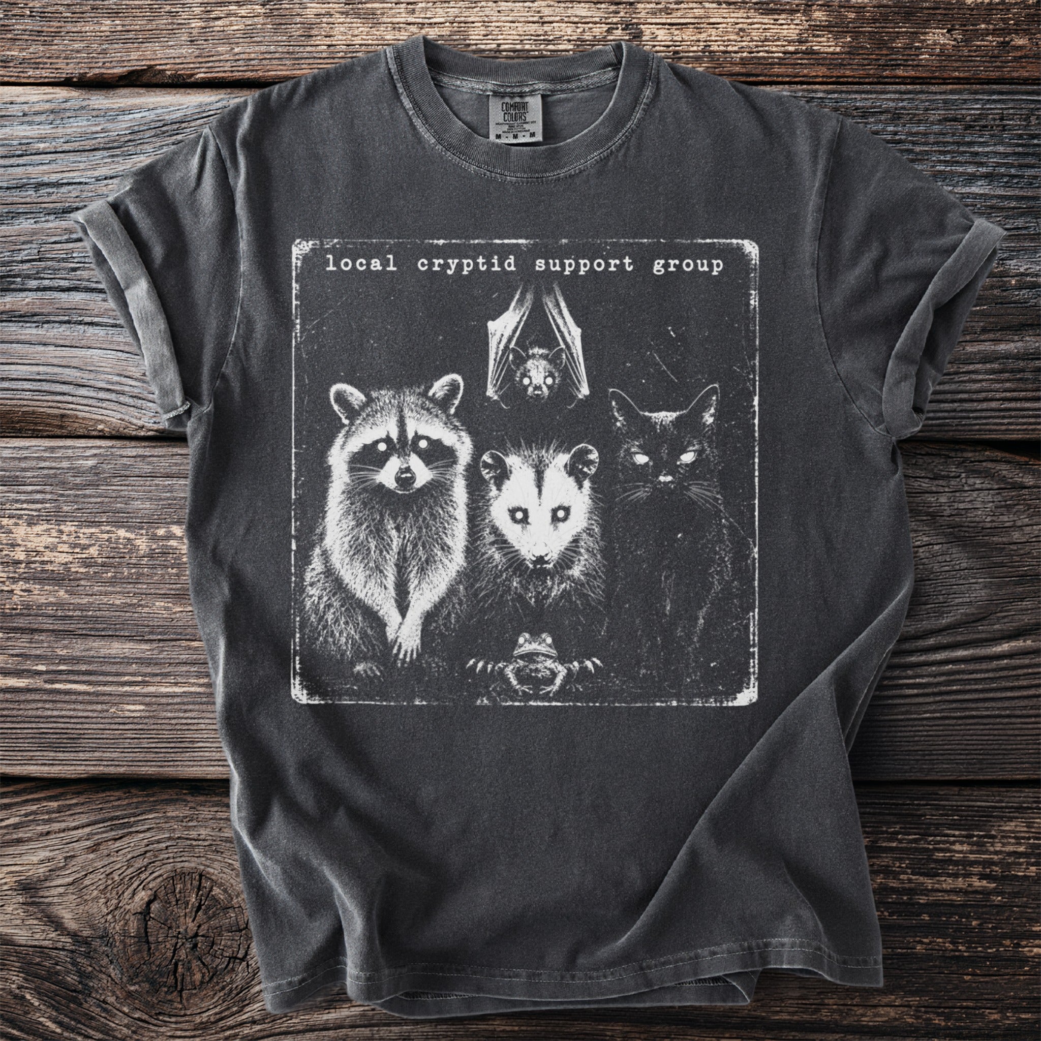 Dark t-shirt with graphic of raccoon, opossum, bat, cat, and frog and text "local cryptid support group".
