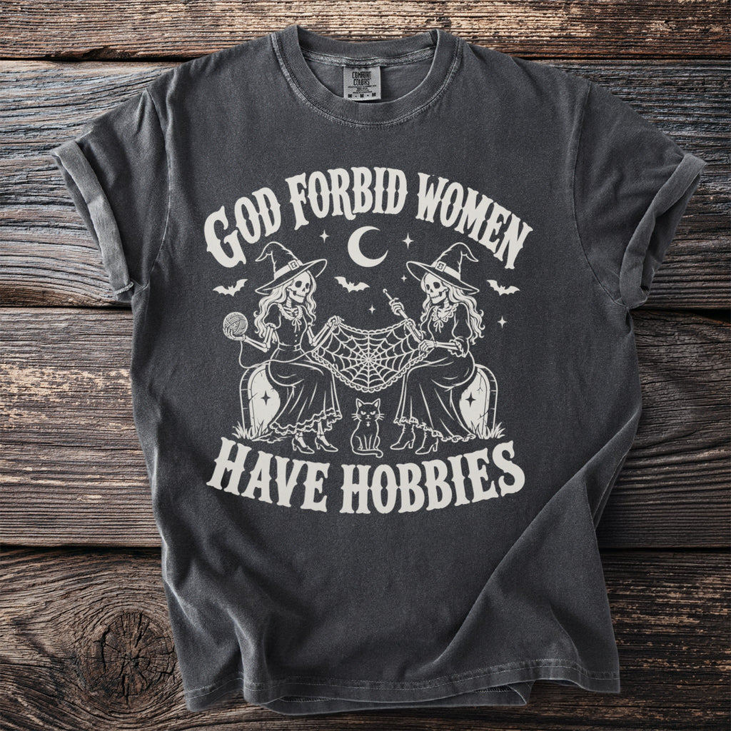 Dark gray t-shirt with graphic of skeletons knitting and crocheting and the words "God Forbid Women Have Hobbies."