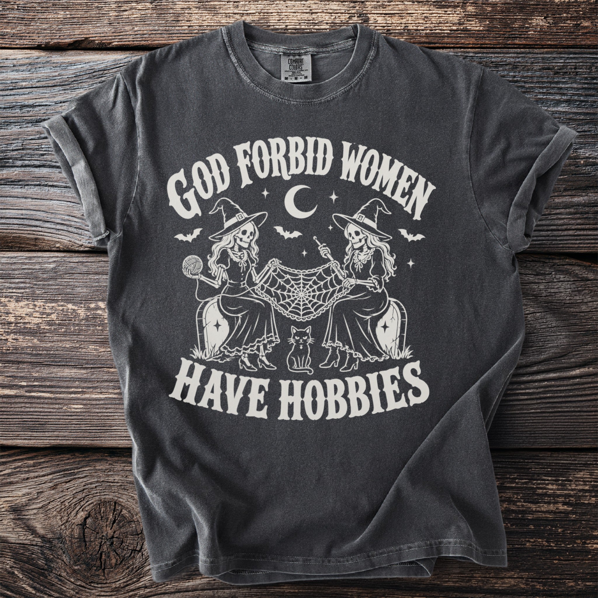 Dark gray t-shirt with graphic of skeletons knitting and crocheting and the words "God Forbid Women Have Hobbies."
