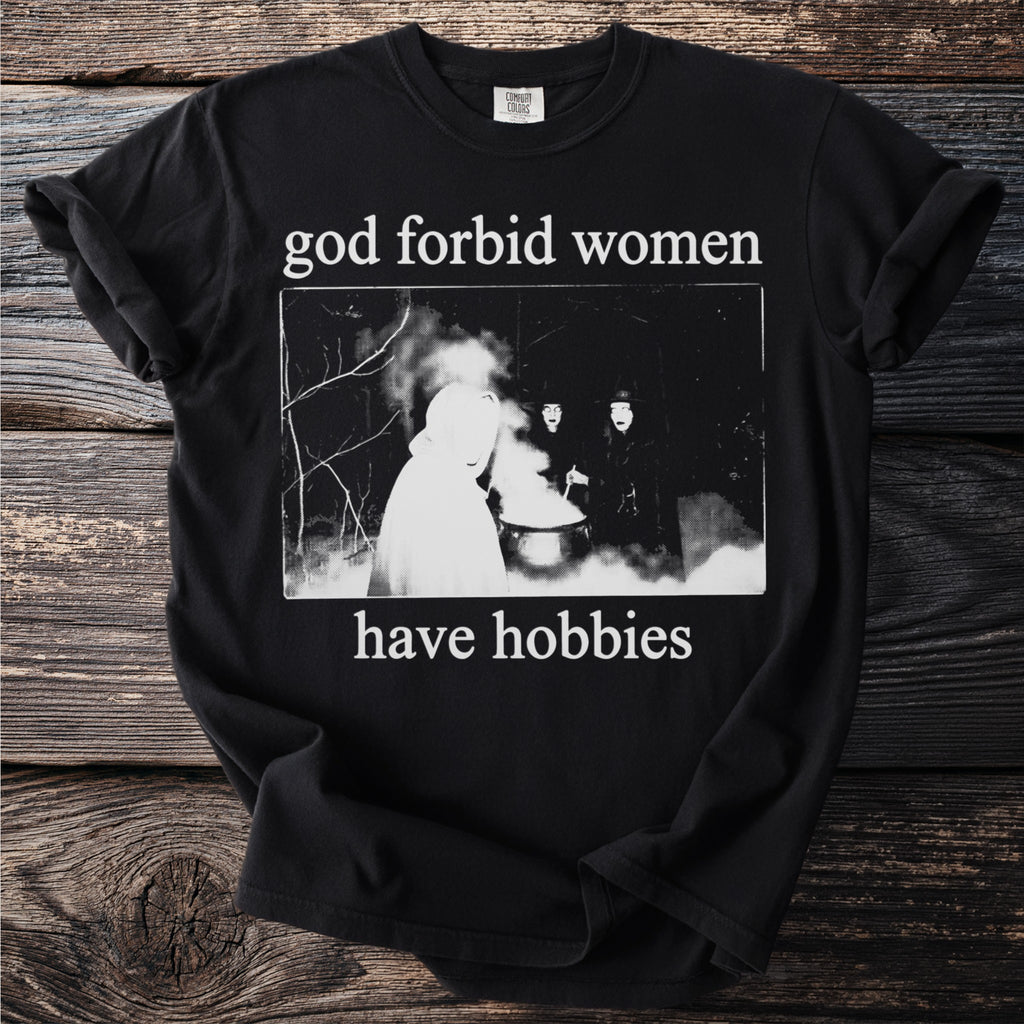 Black t-shirt with white text "god forbid women have hobbies" and a dark illustration.