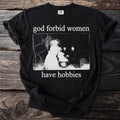 Black t-shirt with white text "god forbid women have hobbies" and a dark illustration.