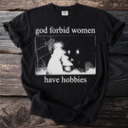 Black t-shirt with white text "god forbid women have hobbies" and a dark illustration.