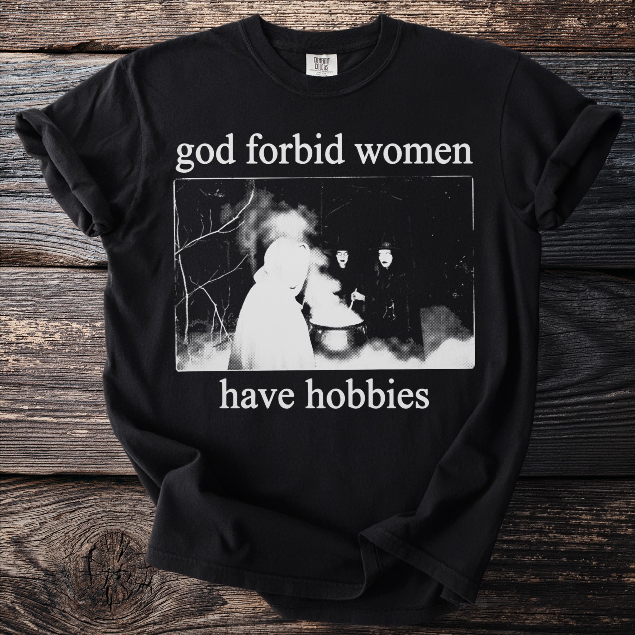 Black t-shirt with white text "god forbid women have hobbies" and a dark illustration.
