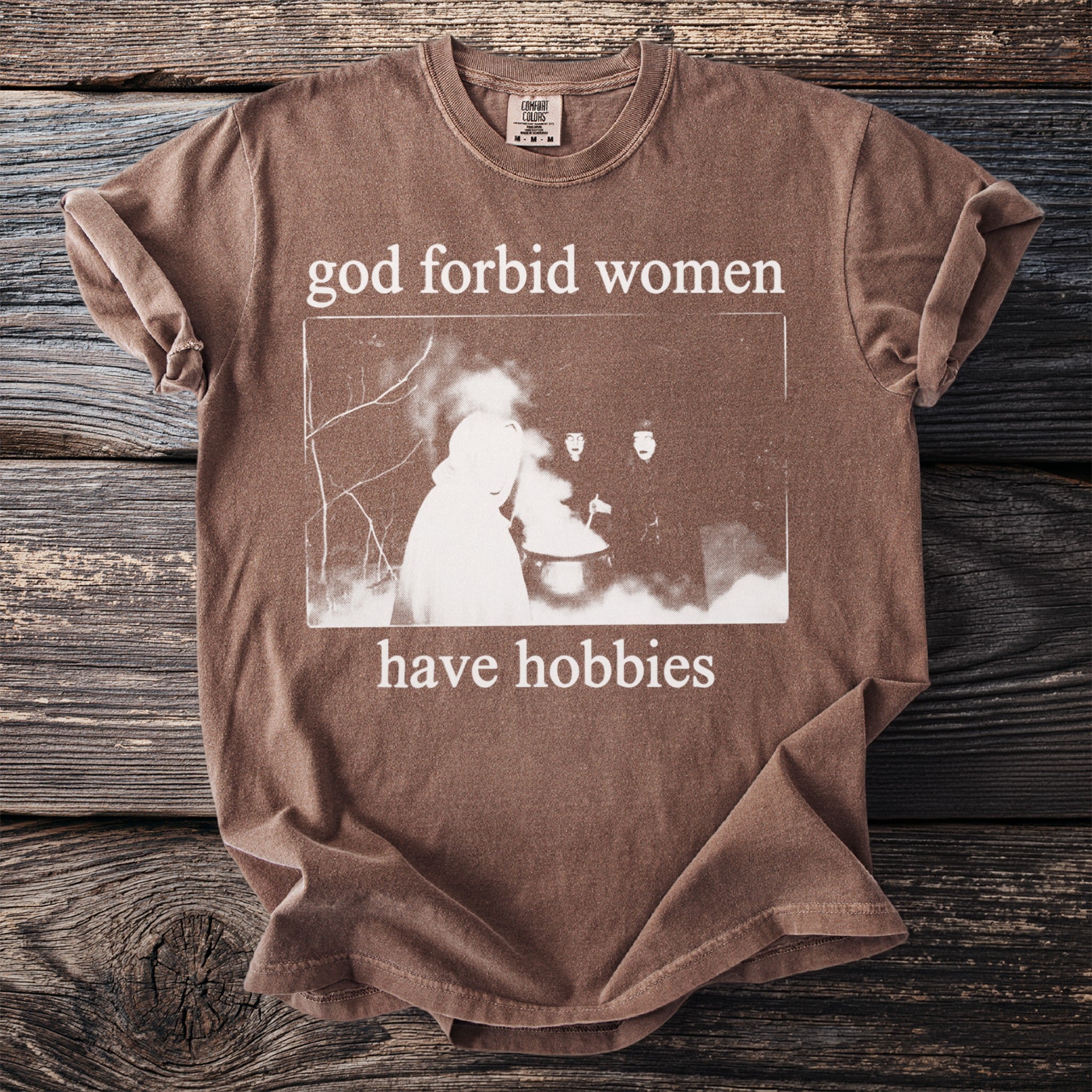 Brown t-shirt with white text "god forbid women have hobbies" and a vintage image.