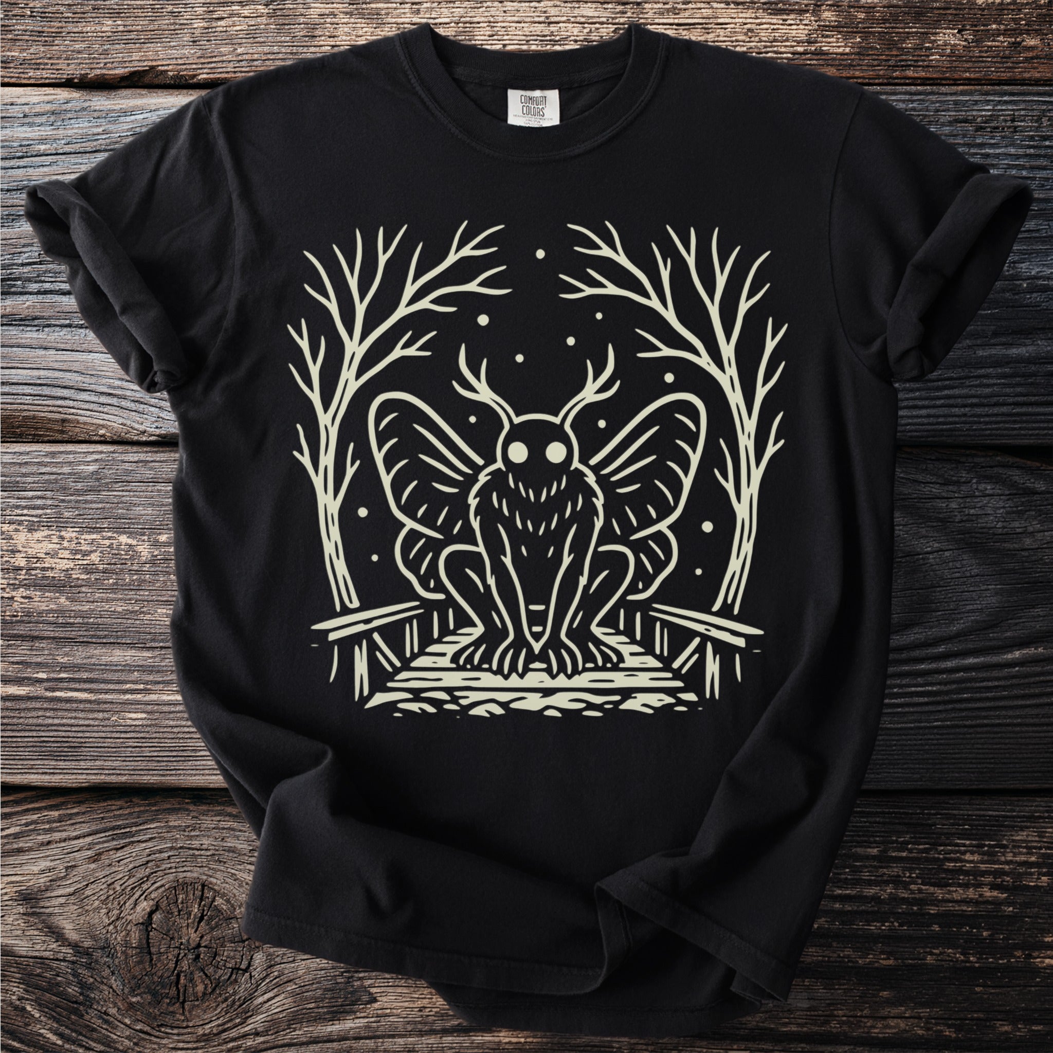 Black t-shirt with cream illustration of a mothman in a forest setting.