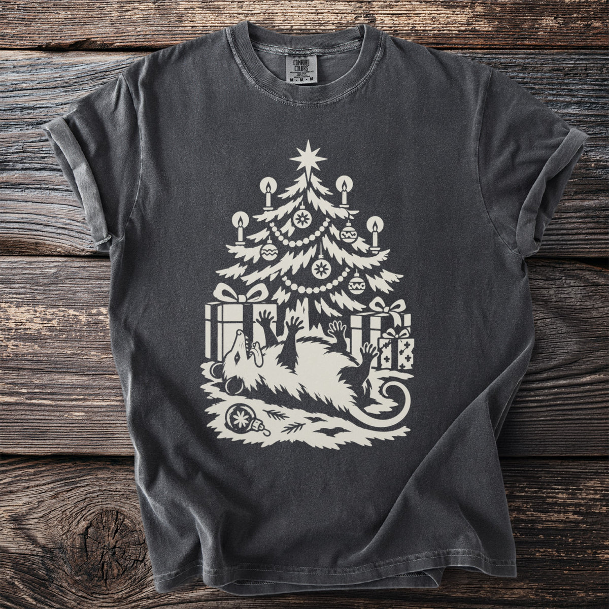 Pepper gray t-shirt with a cream folk-cut print: an opossum lies on its back under a candlelit Christmas tree, playing dead among the presents.