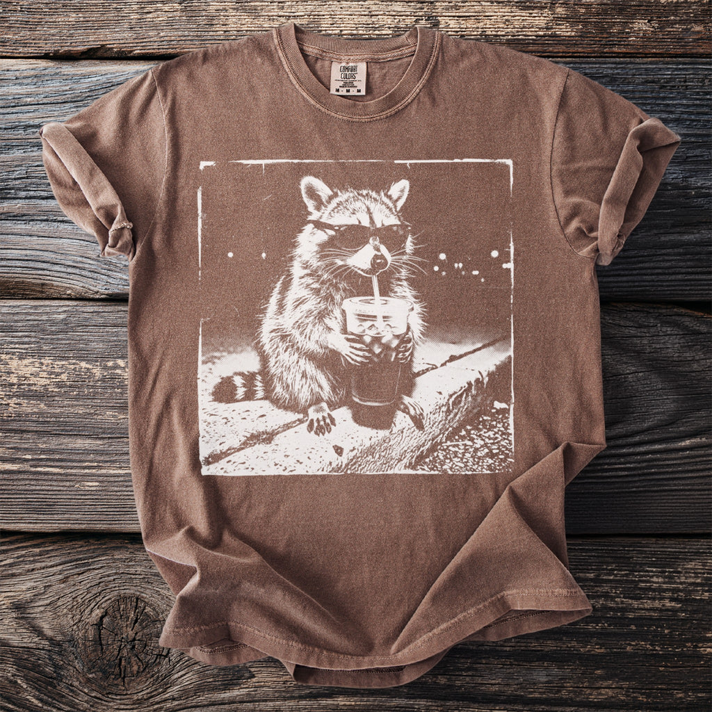 Brown t-shirt with a distressed print of a raccoon wearing sunglasses drinking from a cup.
