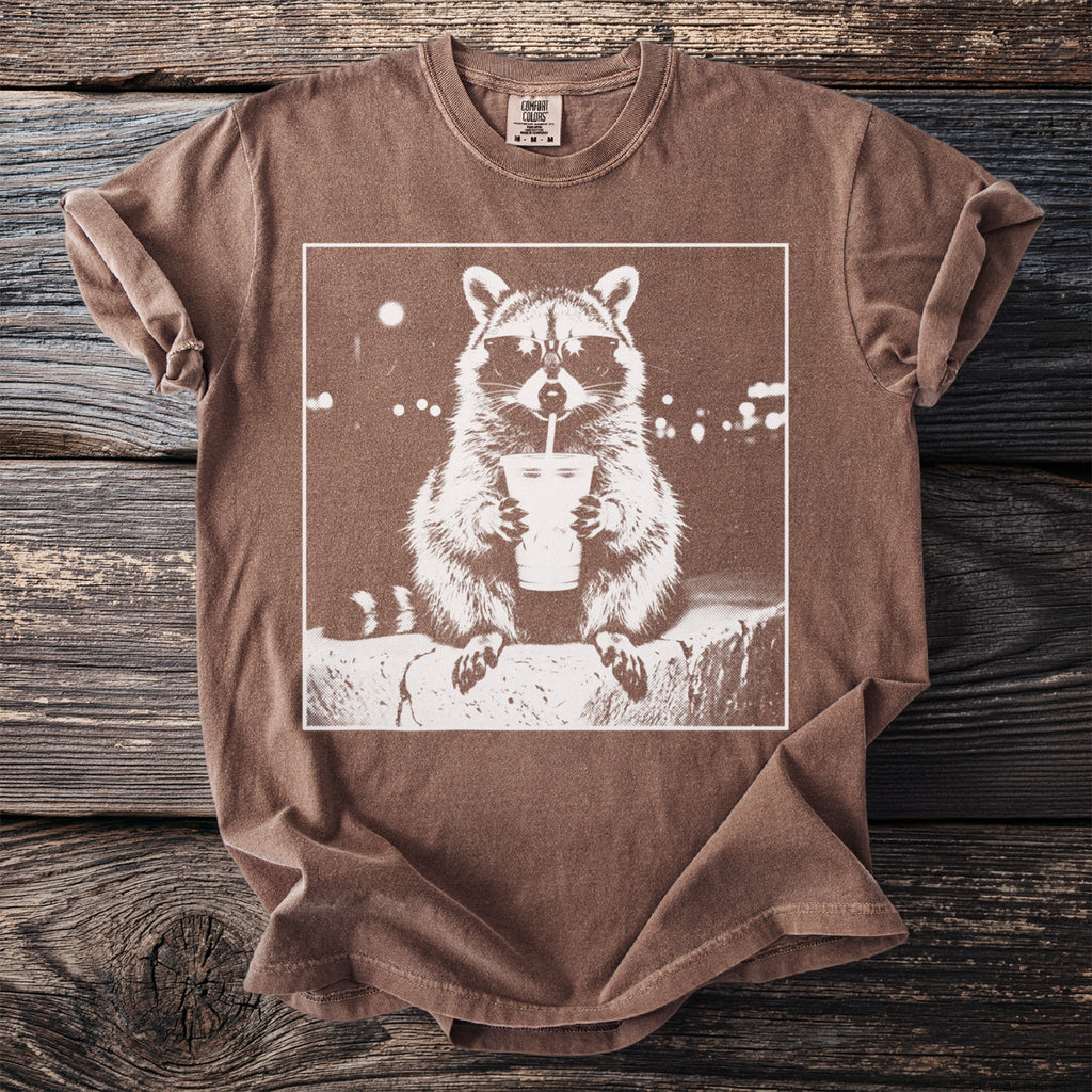 T-shirt with a white graphic of a raccoon wearing sunglasses drinking from a cup.