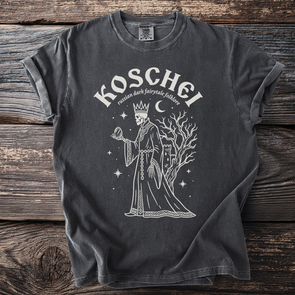 Dark gray t-shirt graphic of Koschei, a skeleton king holding a mirror near a castle and tree.