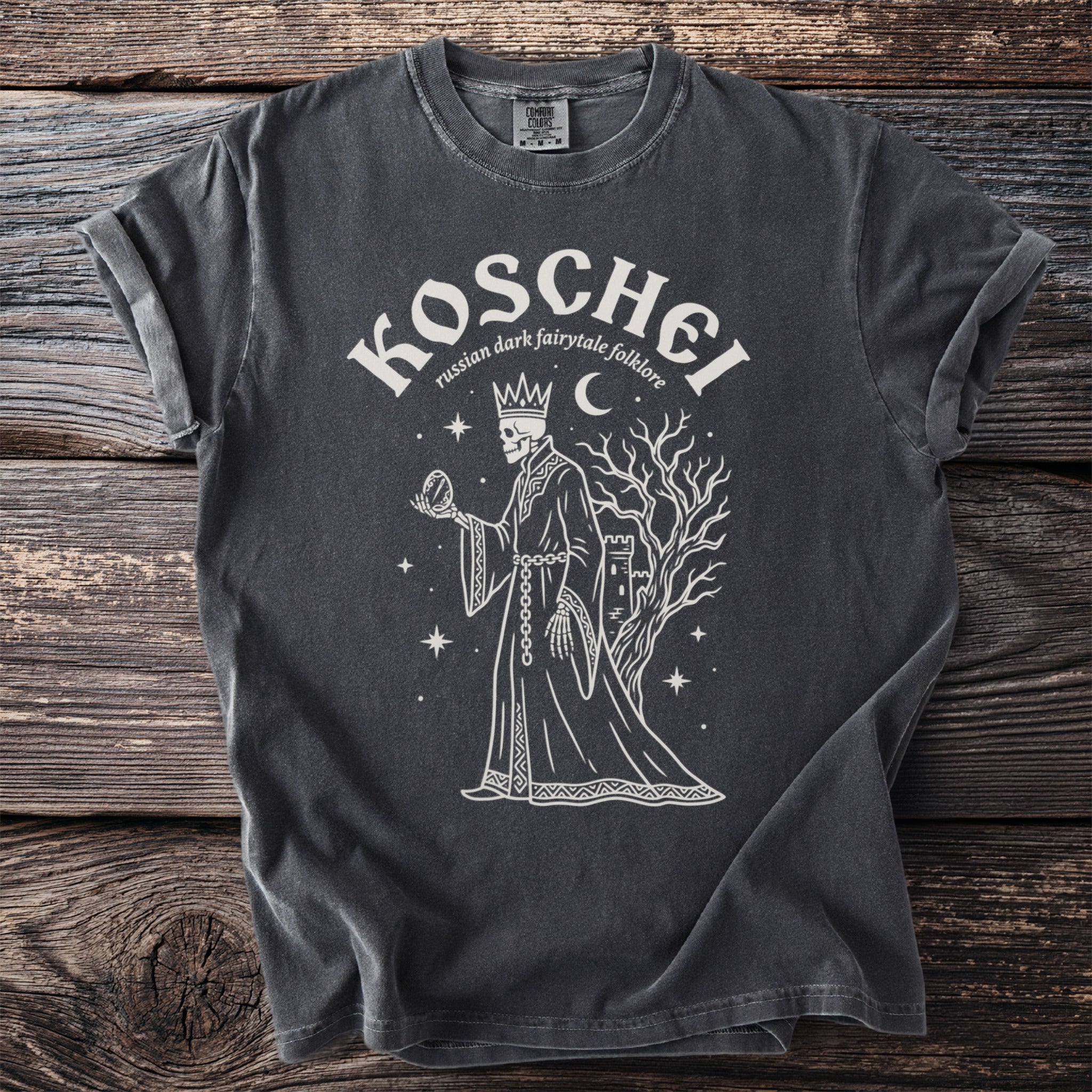 Dark gray t-shirt graphic of Koschei, a skeleton king holding a mirror near a castle and tree.