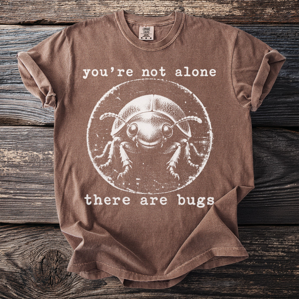 Brown t-shirt with white text "you're not alone" and a round graphic of a smiling beetle, followed by "there are bugs".