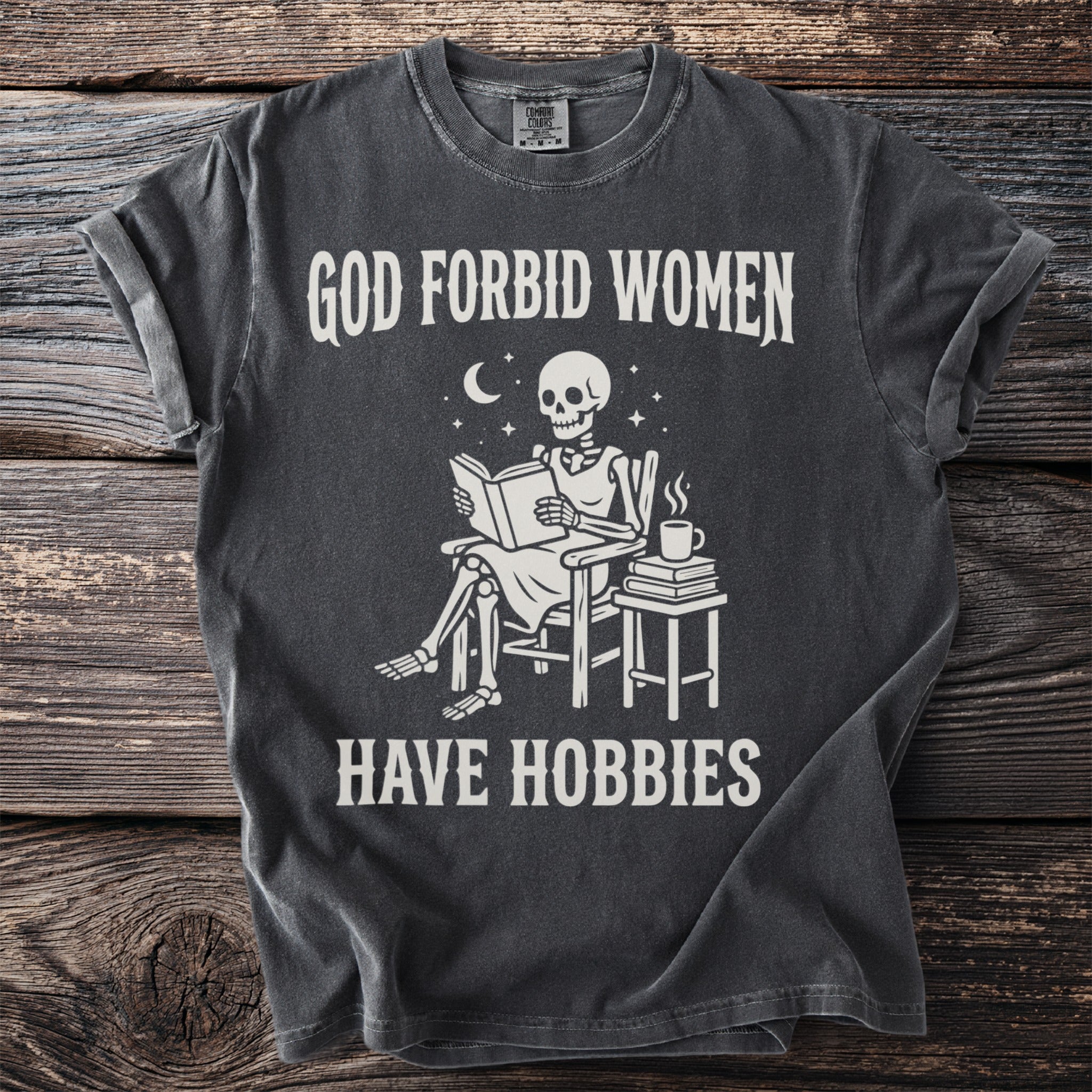 Skeleton sitting in a chair reading with text: God forbid women have hobbies.