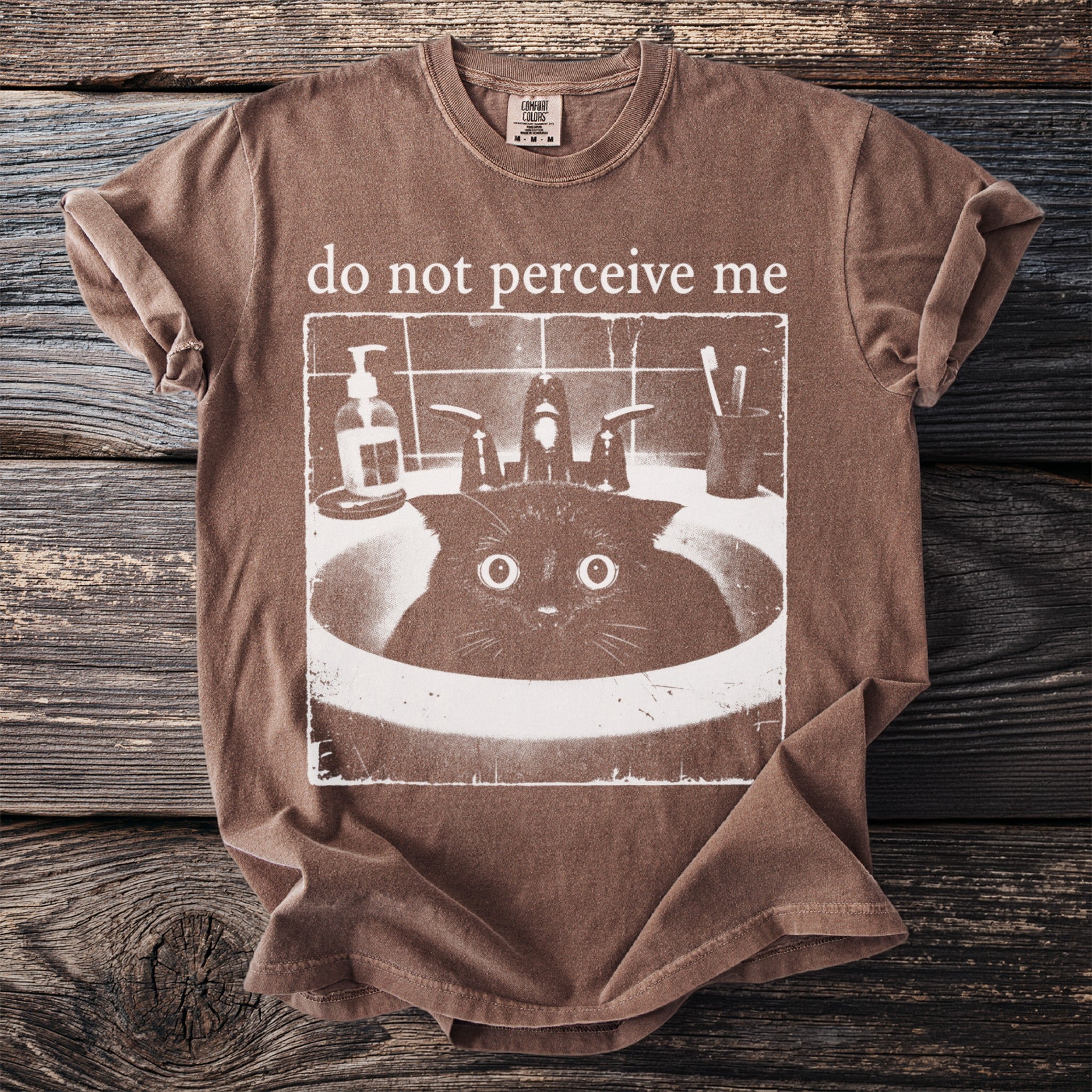 Brown t-shirt with "do not perceive me" text and a cat in a sink graphic.