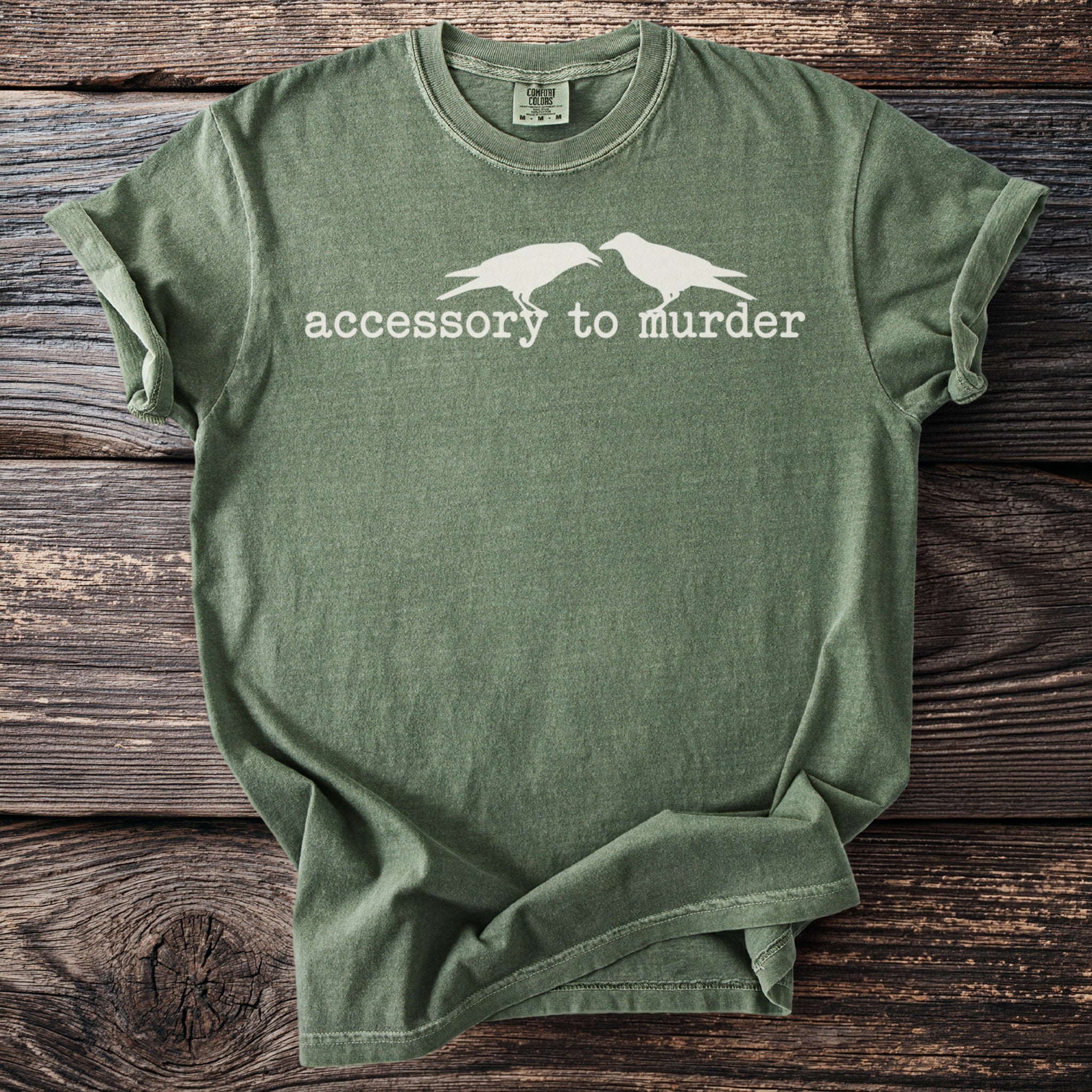 Olive green t-shirt with white crows and text reading "accessory to murder".