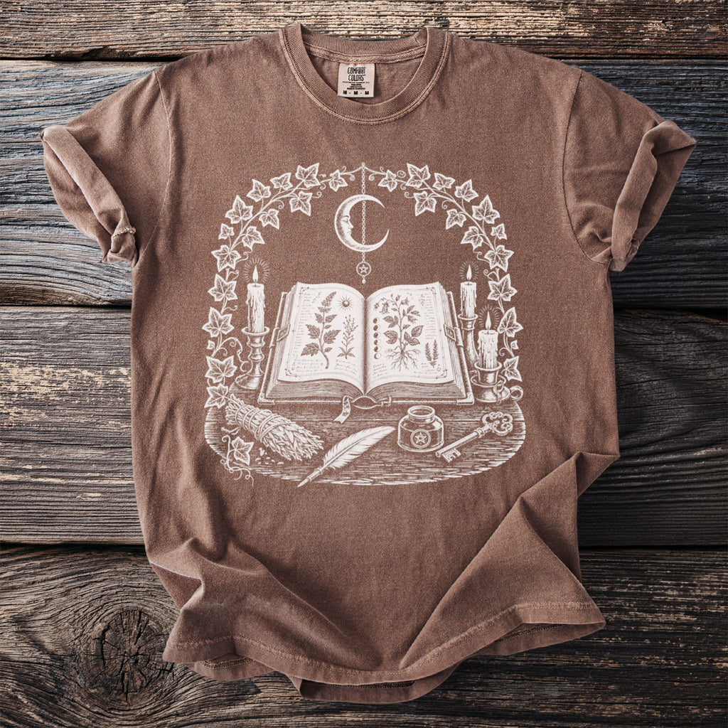 Brown t-shirt with white graphic of an open book, crescent moon, candles, and plants.