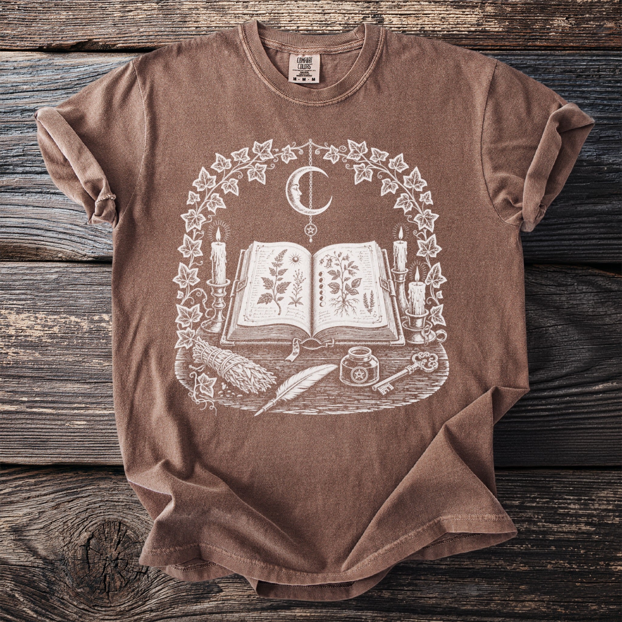 Brown t-shirt with white graphic of an open book, crescent moon, candles, and plants.