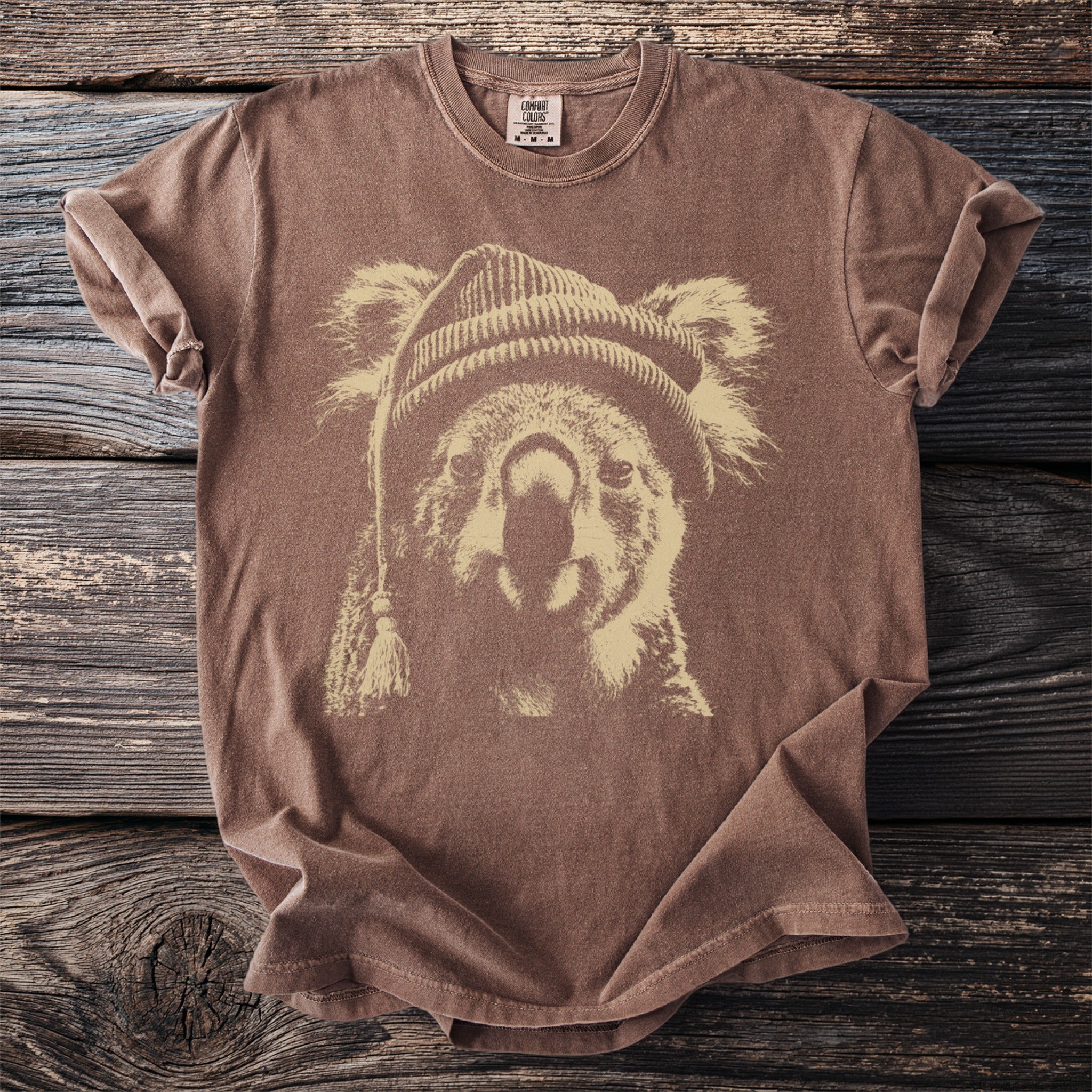 Brown t-shirt with a beige graphic of a koala wearing a beanie on a wooden background.