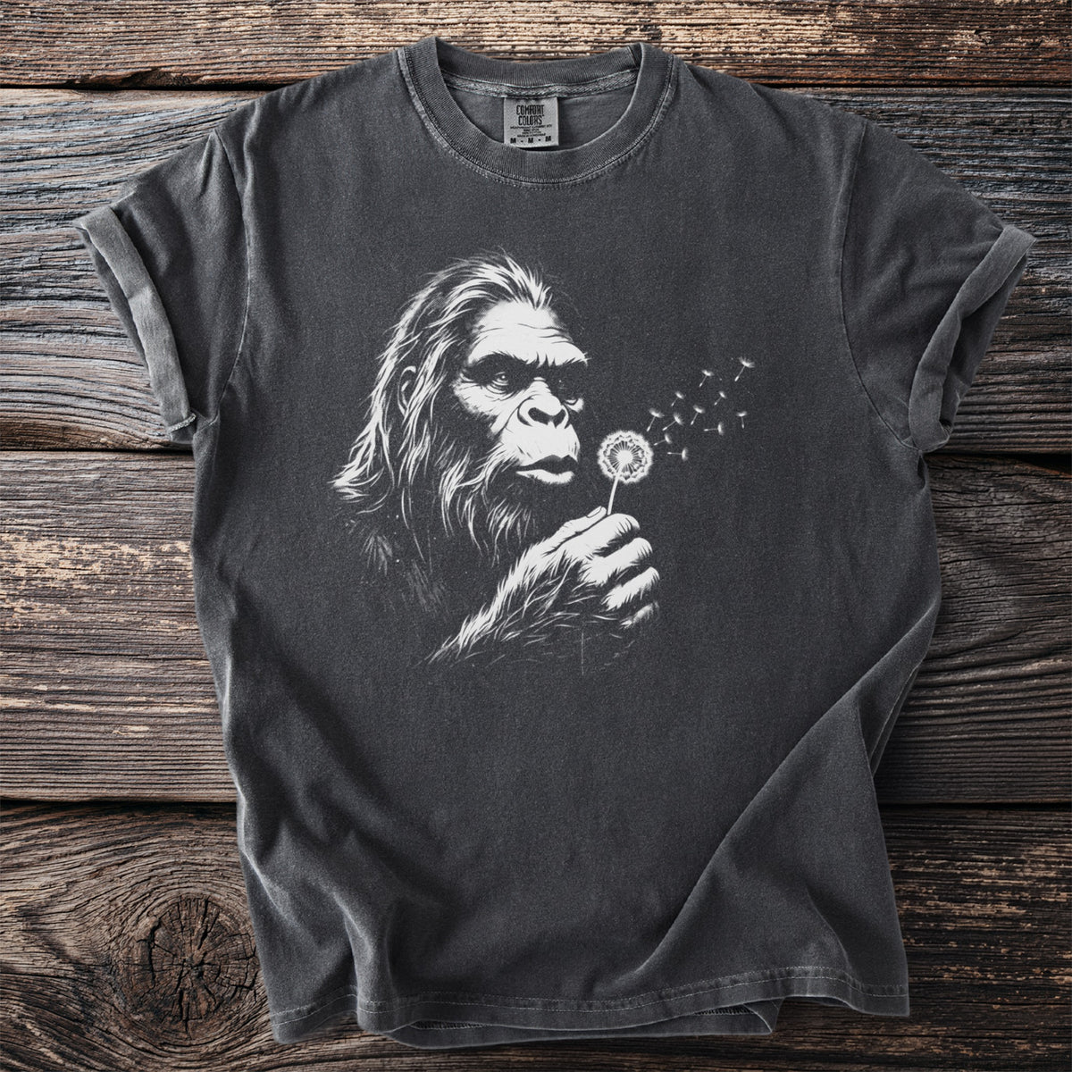 Graphic t-shirt with Bigfoot blowing dandelion seeds on a dark background.