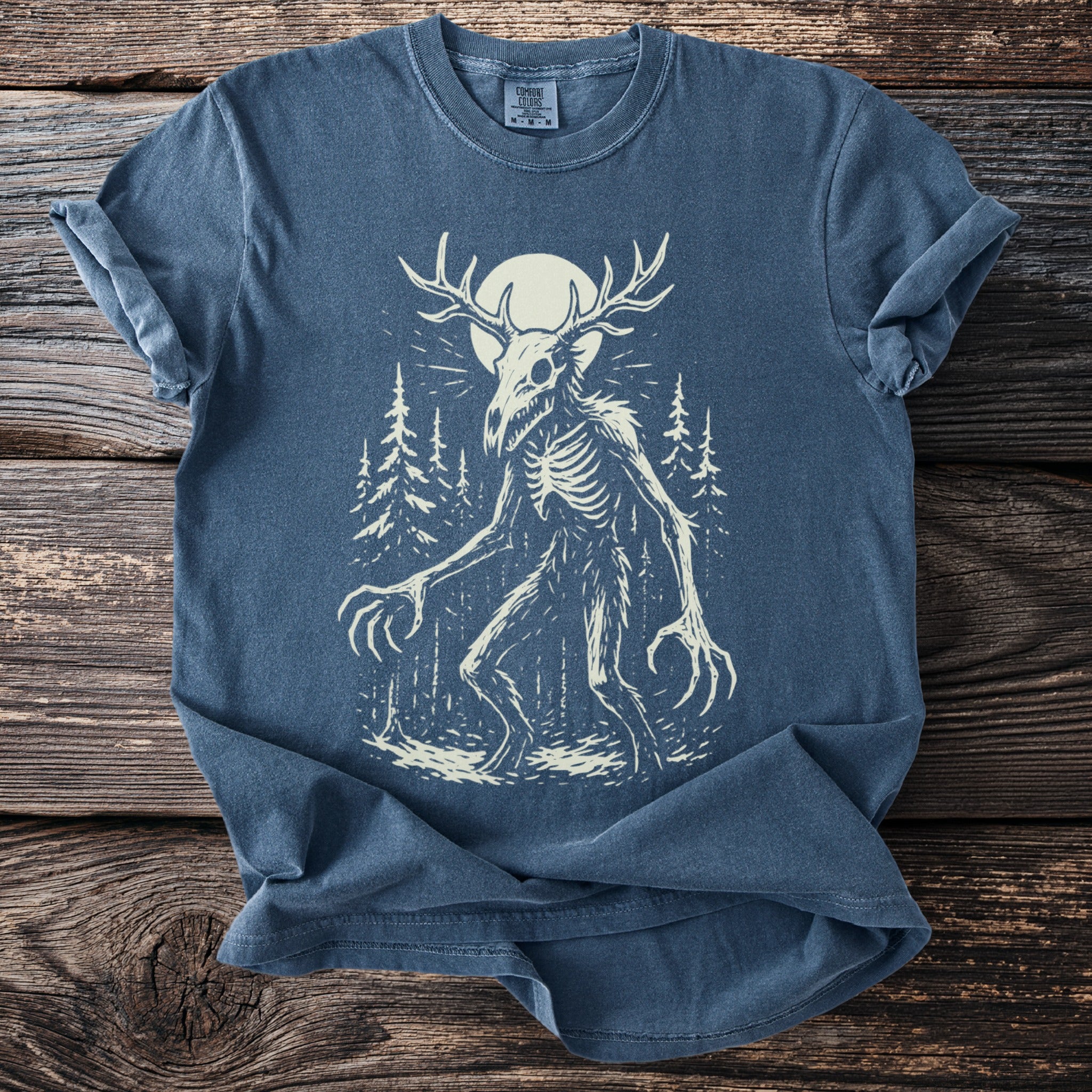 Blue t-shirt with a skeletal creature with antlers and claws in a forest.