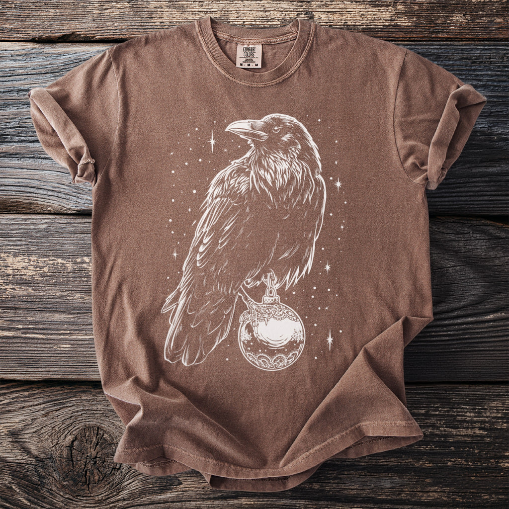 Brown t-shirt with white line art of a crow holding a decorative orb with stars visible.