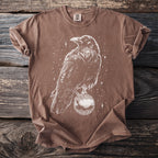 Brown t-shirt with white line art of a crow holding a decorative orb with stars visible.