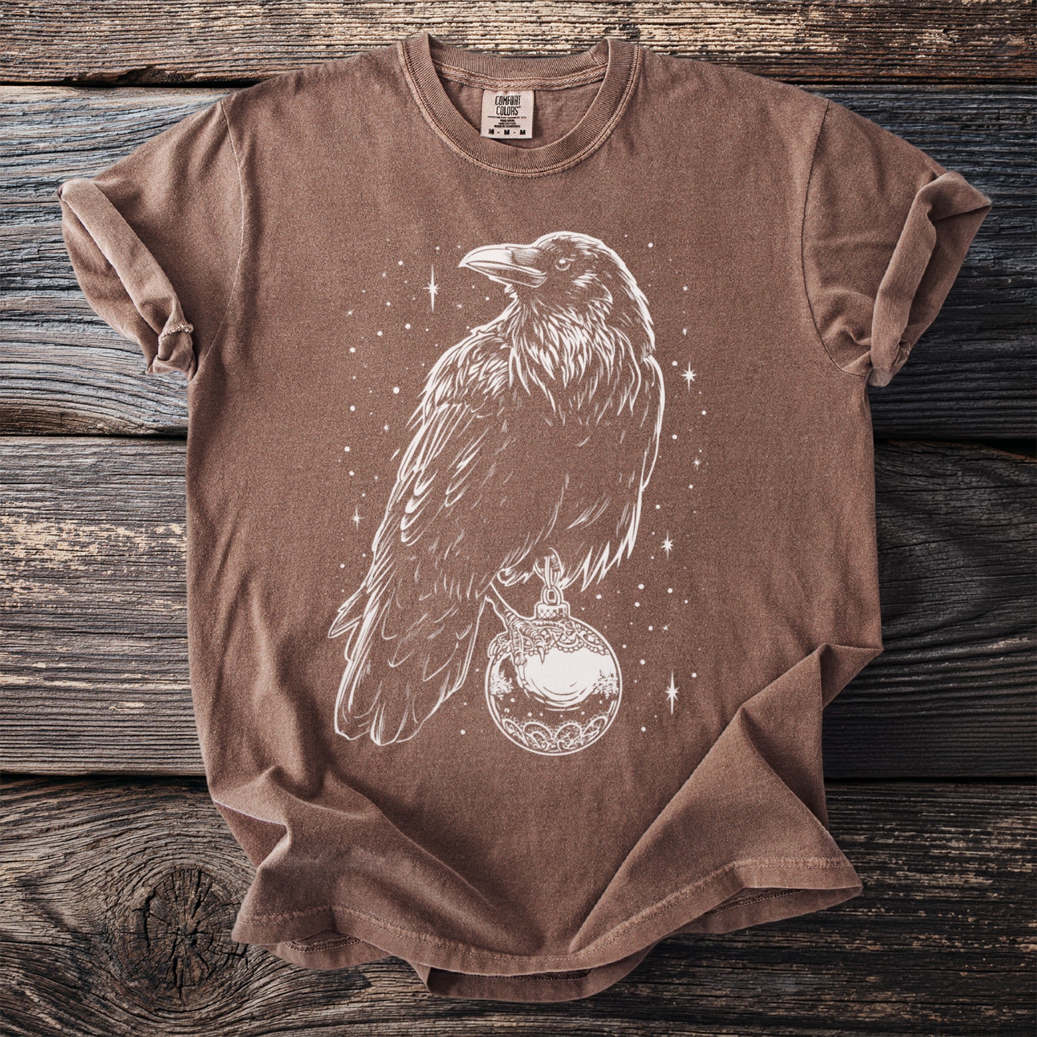 Brown t-shirt with white line art of a crow holding a decorative orb with stars visible.