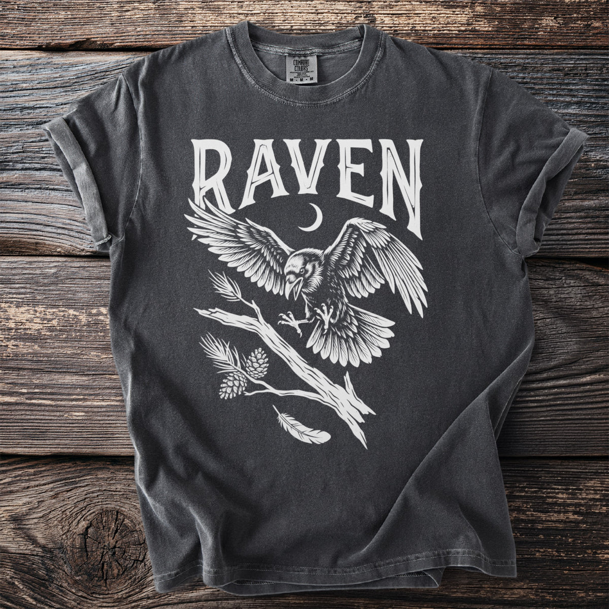 Dark t-shirt with white graphic of a raven in flight above a branch and pinecones.