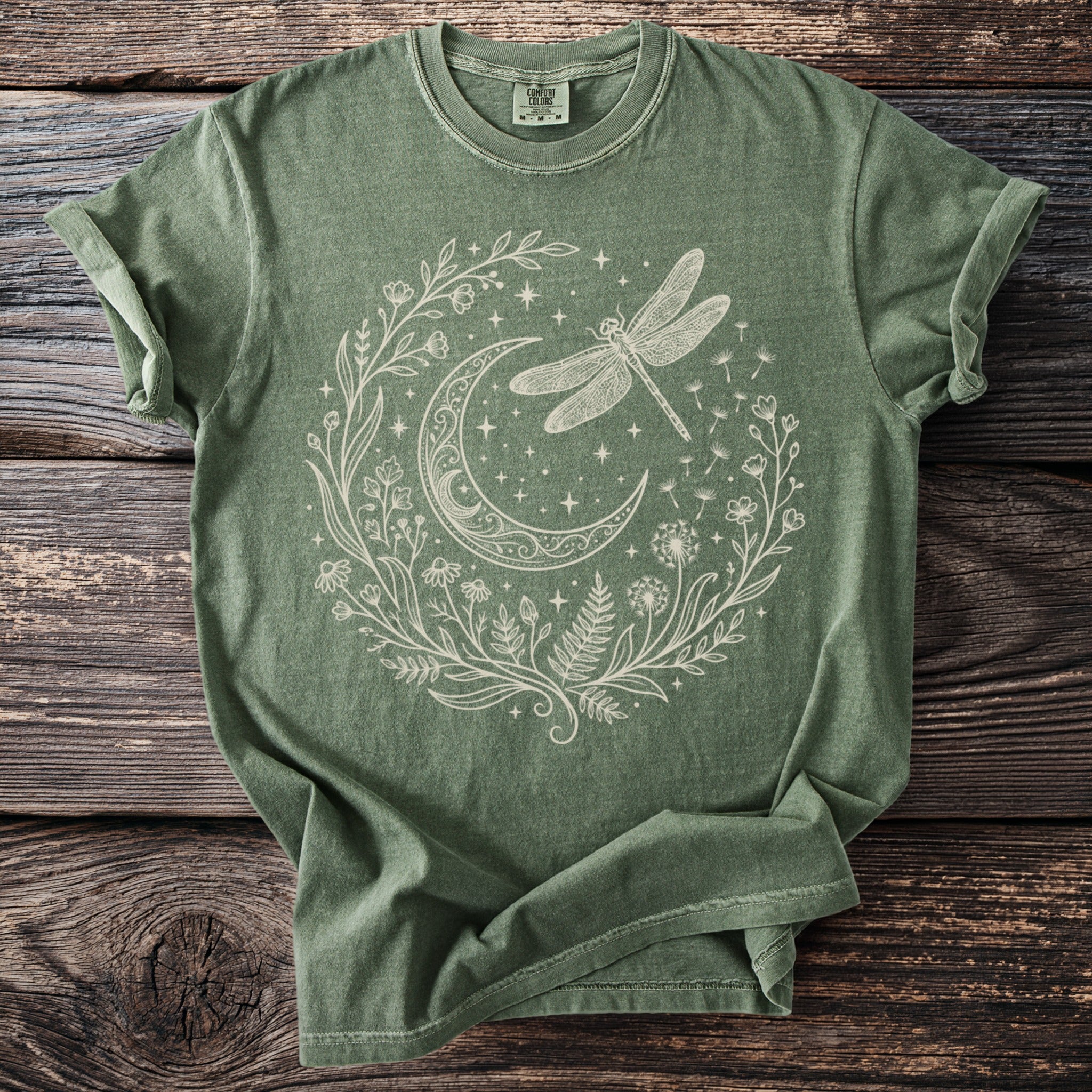 Green t-shirt with cream line art of a dragonfly, moon, and floral wreath.