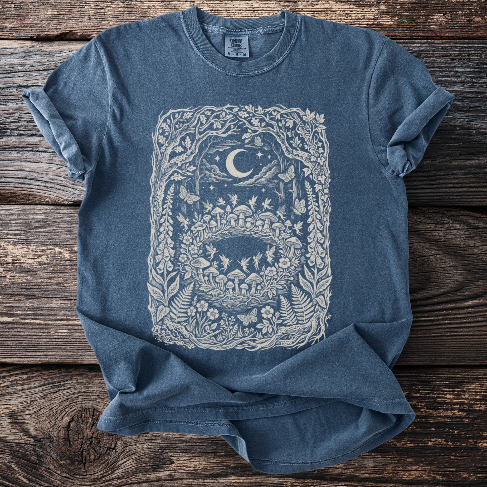 Blue t-shirt with a whimsical forest scene print featuring mushrooms, fairies, and a crescent moon.
