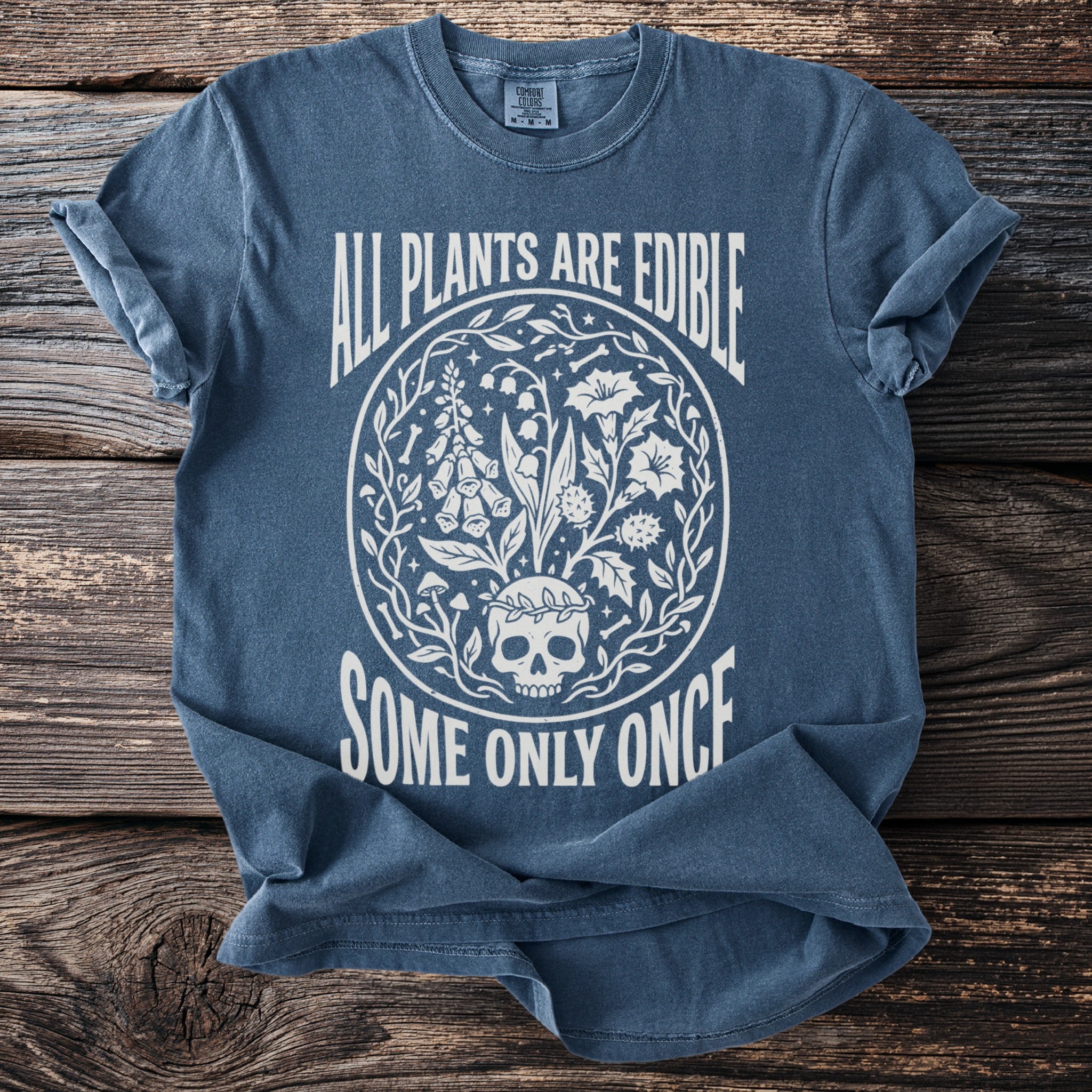 Blue t-shirt with white graphic of a skull, plants, and text "ALL PLANTS ARE EDIBLE SOME ONLY ONCE".
