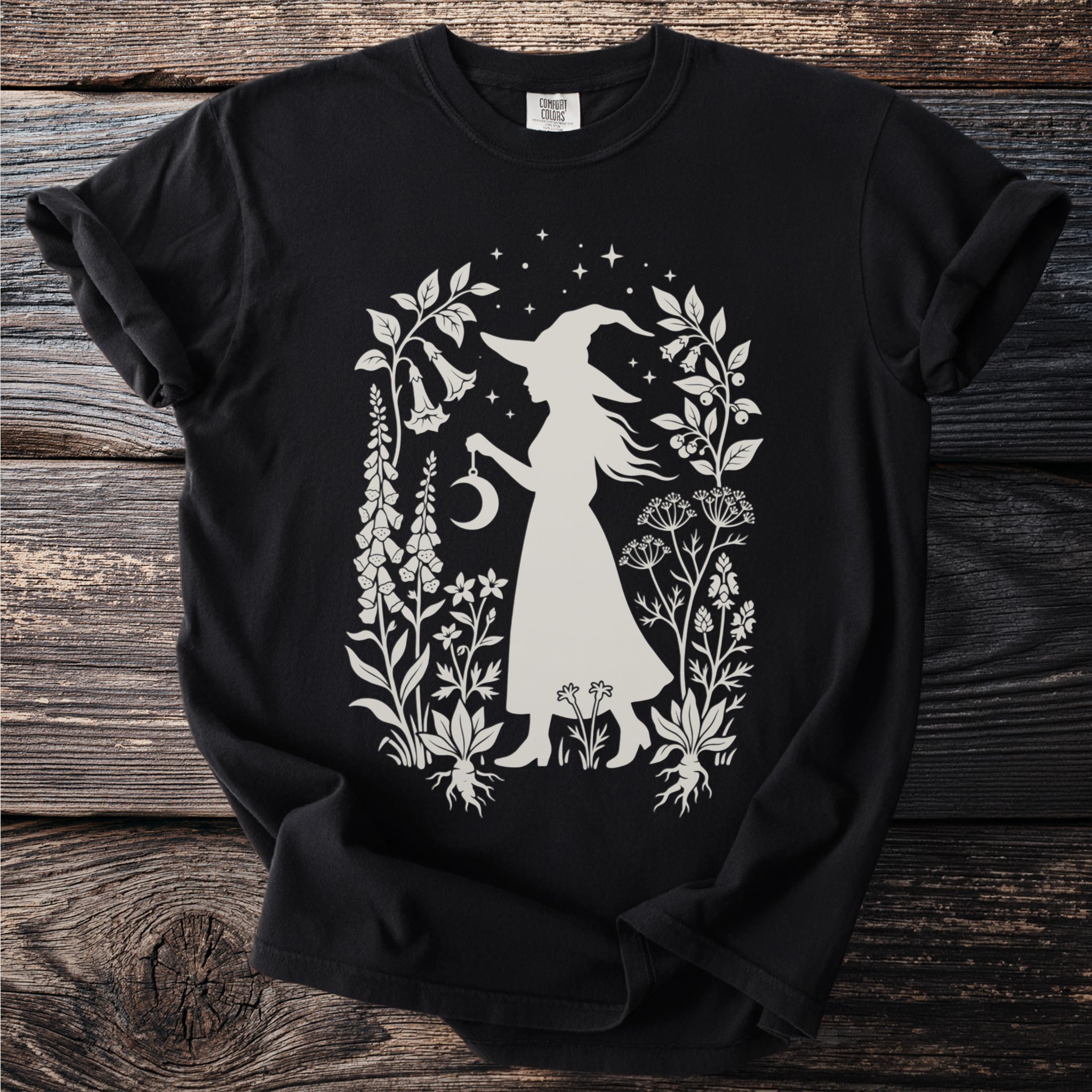 Black t-shirt with a white witch and crescent moon design surrounded by plants and stars.